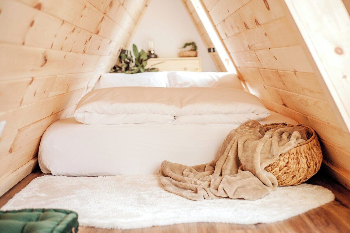 A cozy attic bedroom with a wooden sloped ceiling, a bed with white bedding, a woven basket, and a white rug.