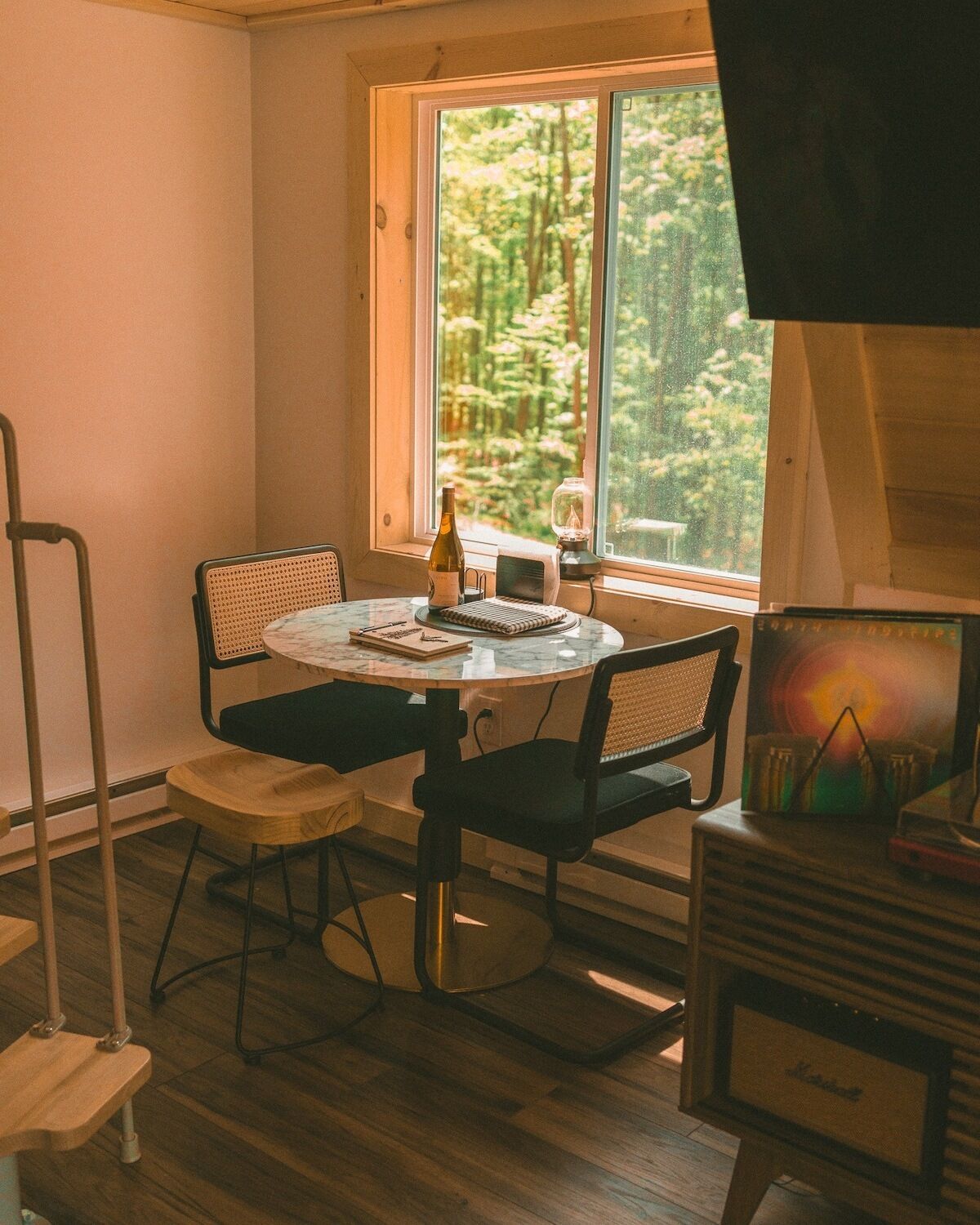 A small, cozy room with a round table, two chairs, a bottle of wine, a small lamp, and a painting on the wall.