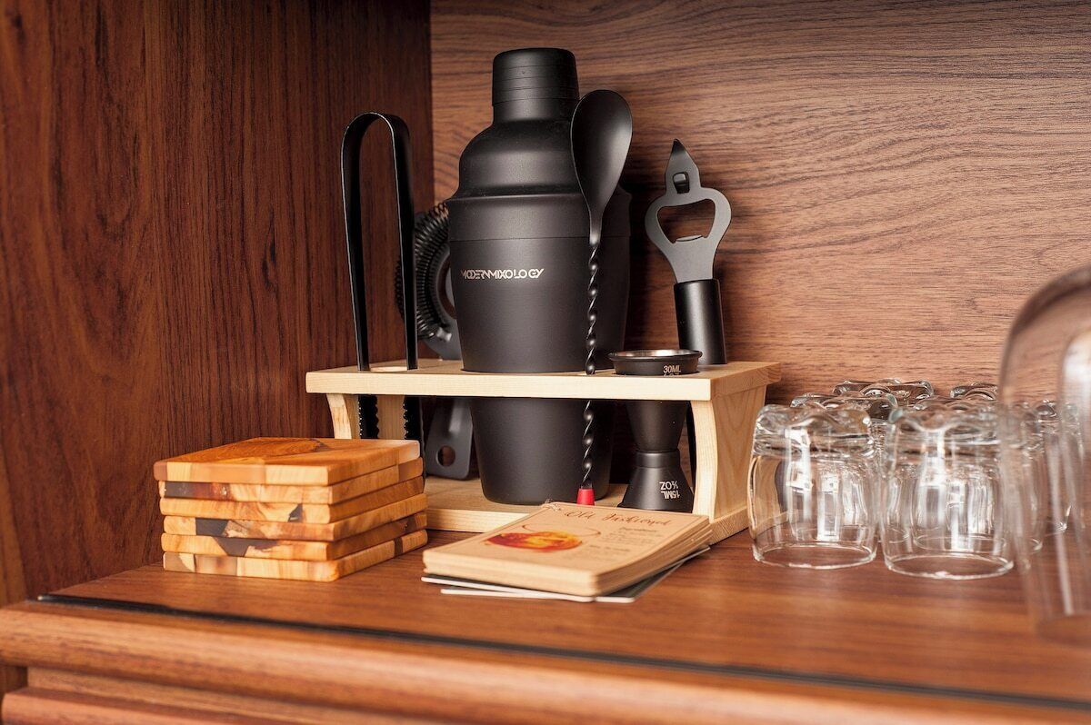 A wooden shelf with a black water bottle, a metal bottle opener, a stack of wooden coasters, a book, and several clear glasses.