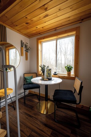 A room with a round table, two chairs, a window with a view of trees, and wooden ceiling beams.