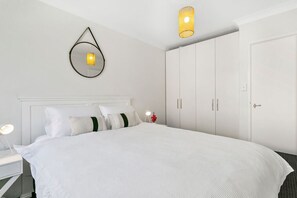 A neatly arranged bedroom with a large bed, a bedside table with a lamp, a wardrobe, and a wall-mounted mirror. - Bondi Bliss Lamrock - 1 minute to beach + Parking (Bondi Beach)