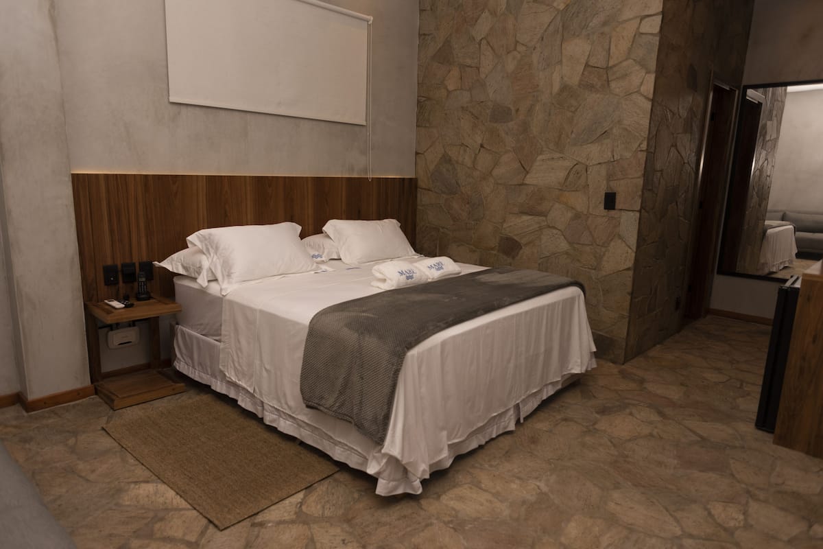 A hotel room with a stone wall, a wooden headboard, a bed with white bedding, a bedside table with a lamp, and a small table with a tablecloth.