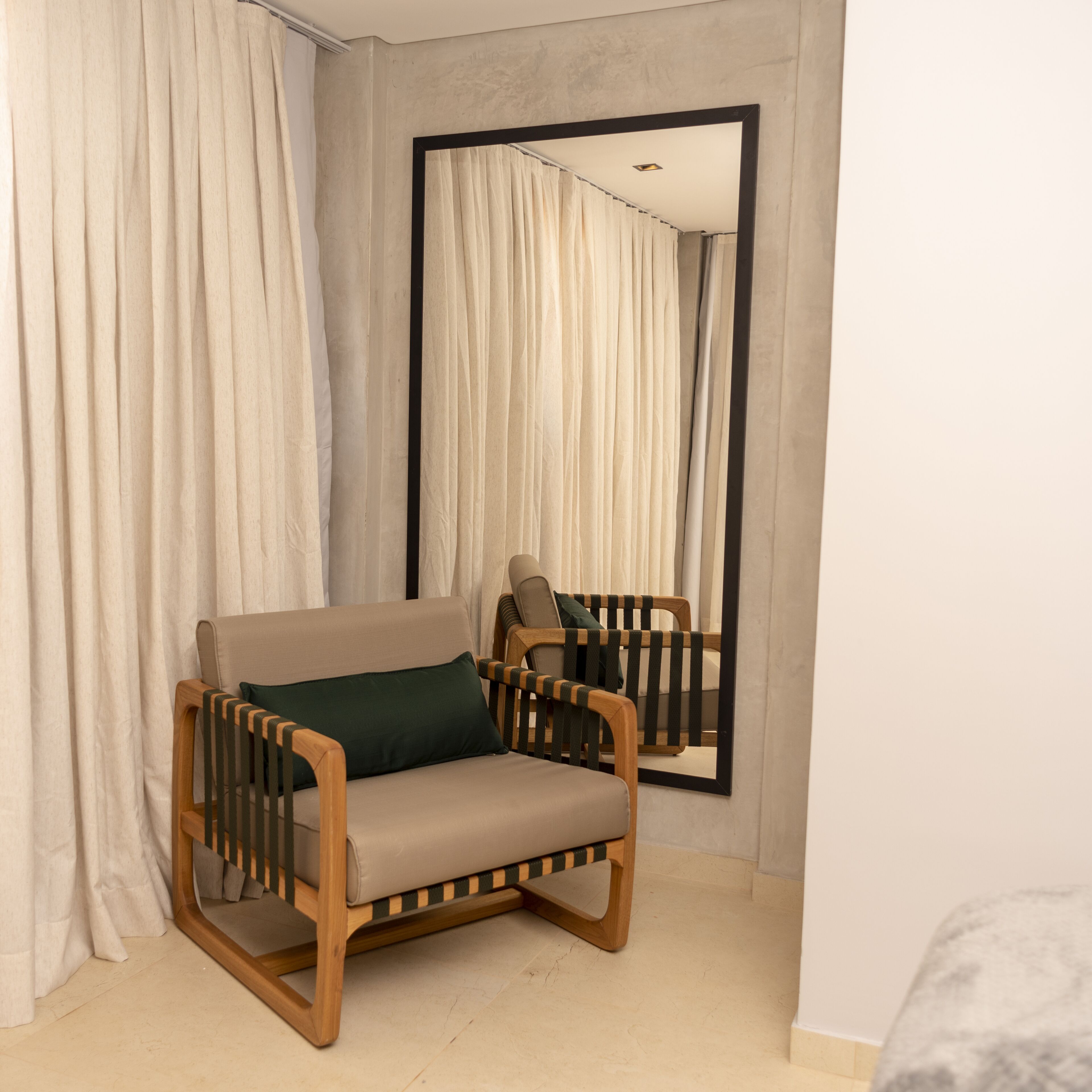 A room with a wooden-framed chair, beige curtains, and a large mirror on the wall.