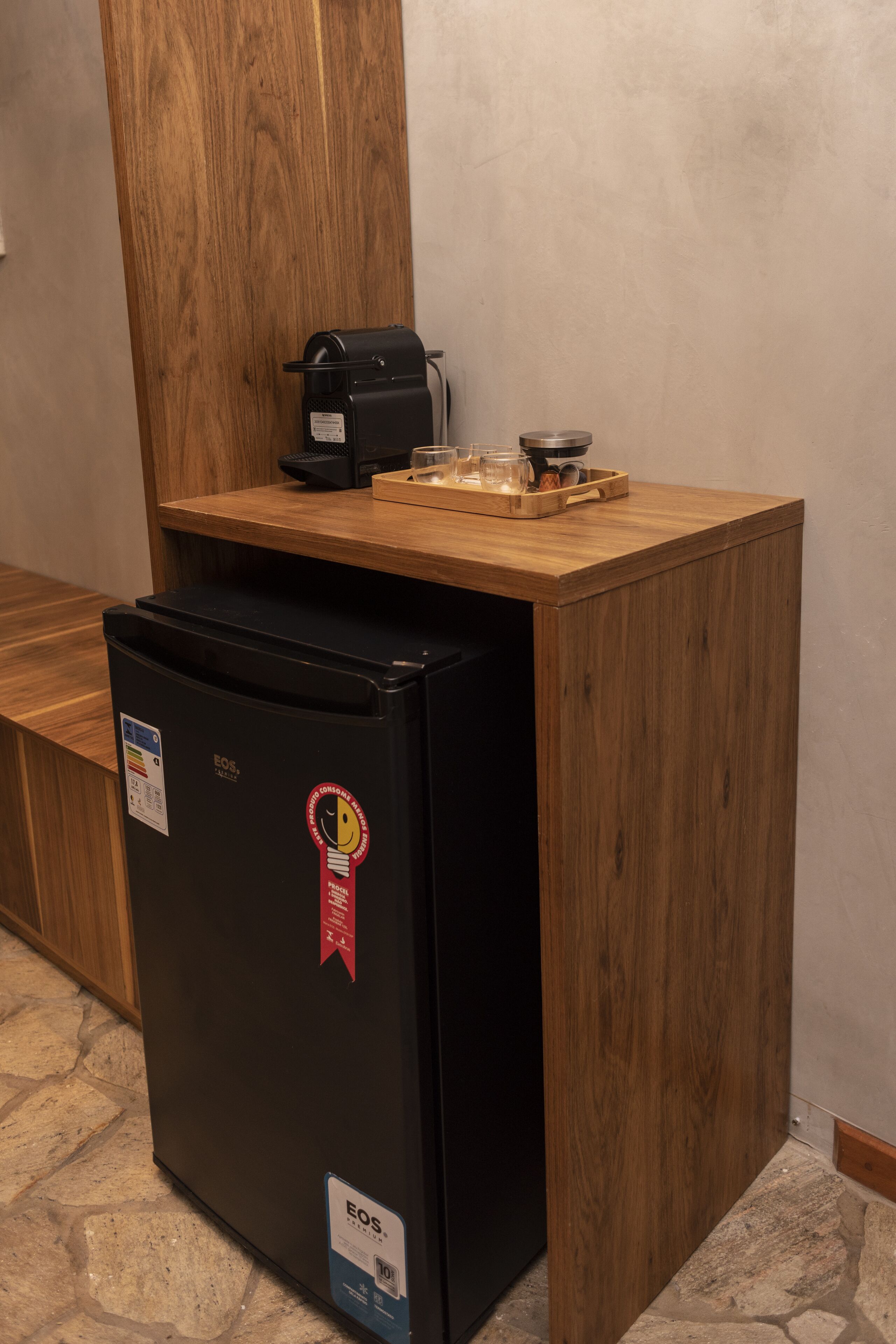 Eine kompakte Küchenzeile mit einem schwarzen Kühlschrank, einer Kaffeemaschine und einer Ablage mit einer Tasse und einem kleinen Behälter.