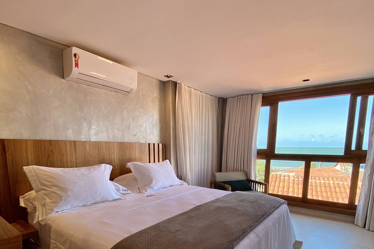 A hotel room with a large bed, a wooden headboard, a view of the sea, and an air conditioning unit on the wall.