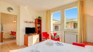 A hotel room with a bed, a TV, a desk, a bookshelf, a red chair, and a window with curtains.
