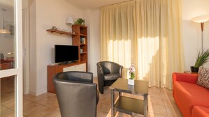 A living room with a TV, two chairs, a sofa, a table, a bookshelf, and a window with curtains.