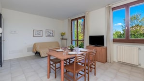 A dining area with a table set for four, a sofa, a TV, and a window with a view of greenery. - Porto Letizia Verdi 5, Porlezza, Italy (Porlezza)