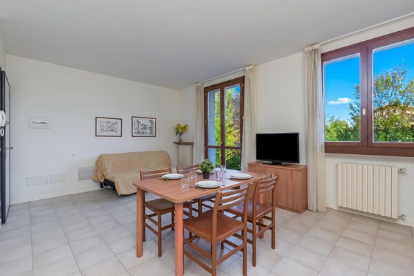 A dining area with a table set for four, a sofa, a TV, and a window with a view of greenery. - Porto Letizia Verdi 5, Porlezza, Italy (Porlezza)