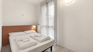 A neatly made bed with white linens and towels, a wooden headboard, a wall-mounted light, and sheer curtains on a window. - Porto Letizia Mozart 5, Porlezza, Italy (Porlezza)