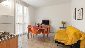 A modern kitchen with a gas stove, dining table with chairs, a TV, and a yellow sofa in a bright living area. - Porto Letizia Mozart 5, Porlezza, Italy (Porlezza)