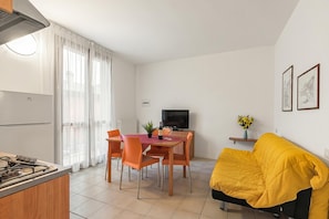 Una cucina moderna con piano cottura a gas, tavolo da pranzo con sedie, televisore e un divano giallo in un soggiorno luminoso.