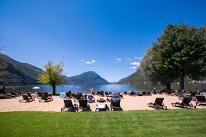 A lakeside beach with lounge chairs and tables, surrounded by mountains and clear skies. - Porto Letizia Mozart 4, Porlezza, Italy (Porlezza)