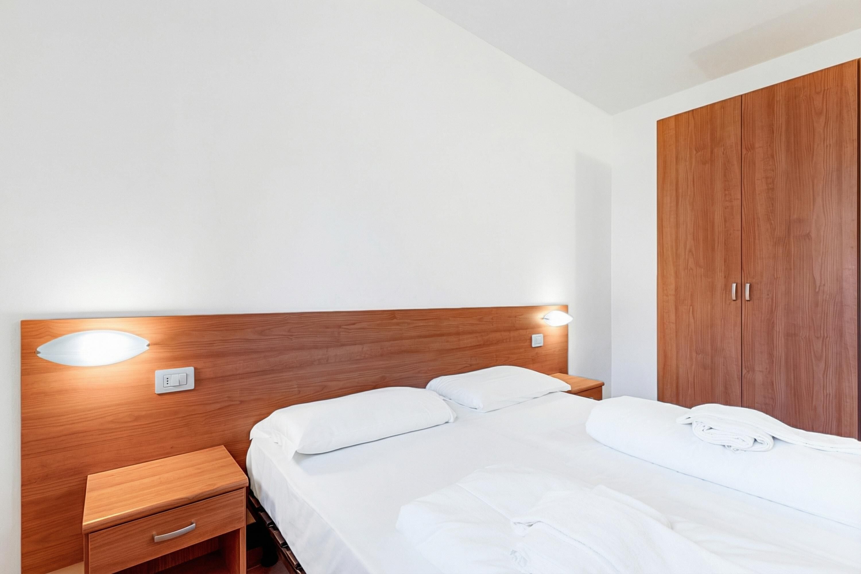 Une chambre d’hôtel avec une tête de lit en bois, un lit simple avec une literie blanche, une table de chevet avec un tiroir et une armoire en bois.