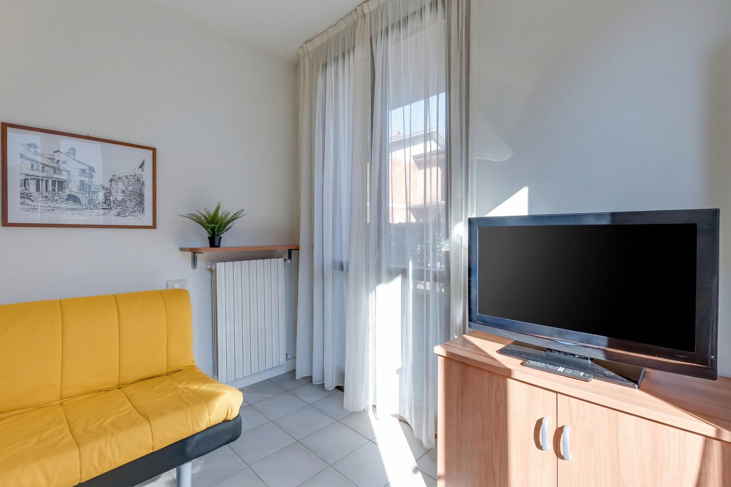 A living room with a yellow sofa, a television on a wooden cabinet, a framed picture on the wall, and a window with sheer curtains.