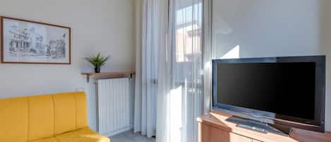 A living room with a yellow sofa, a television on a wooden cabinet, a framed picture on the wall, and a window with sheer curtains.