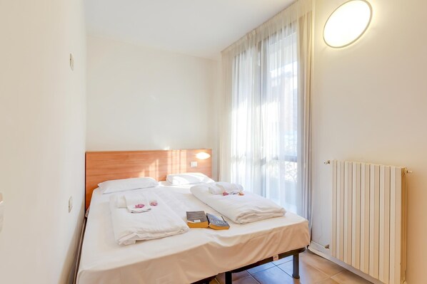 A hotel room with two beds, a wooden headboard, a bedside table, a radiator, and a window with curtains.