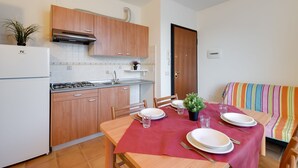A compact kitchen with wooden cabinets, a gas stove, and a sink. A dining table with a red tablecloth is set with plates, glasses, and cutlery. A striped sofa is in the background. - Porto Letizia Chopin 9, Porlezza, Italy (Porlezza)