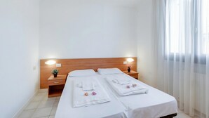 Quarto de hotel com duas camas, cabeceira de madeira, luminárias laterais, uma pequena planta e uma janela com cortinas finas.