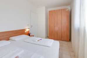 A hotel room with two beds, a wooden wardrobe, a nightstand with a lamp, and a door.