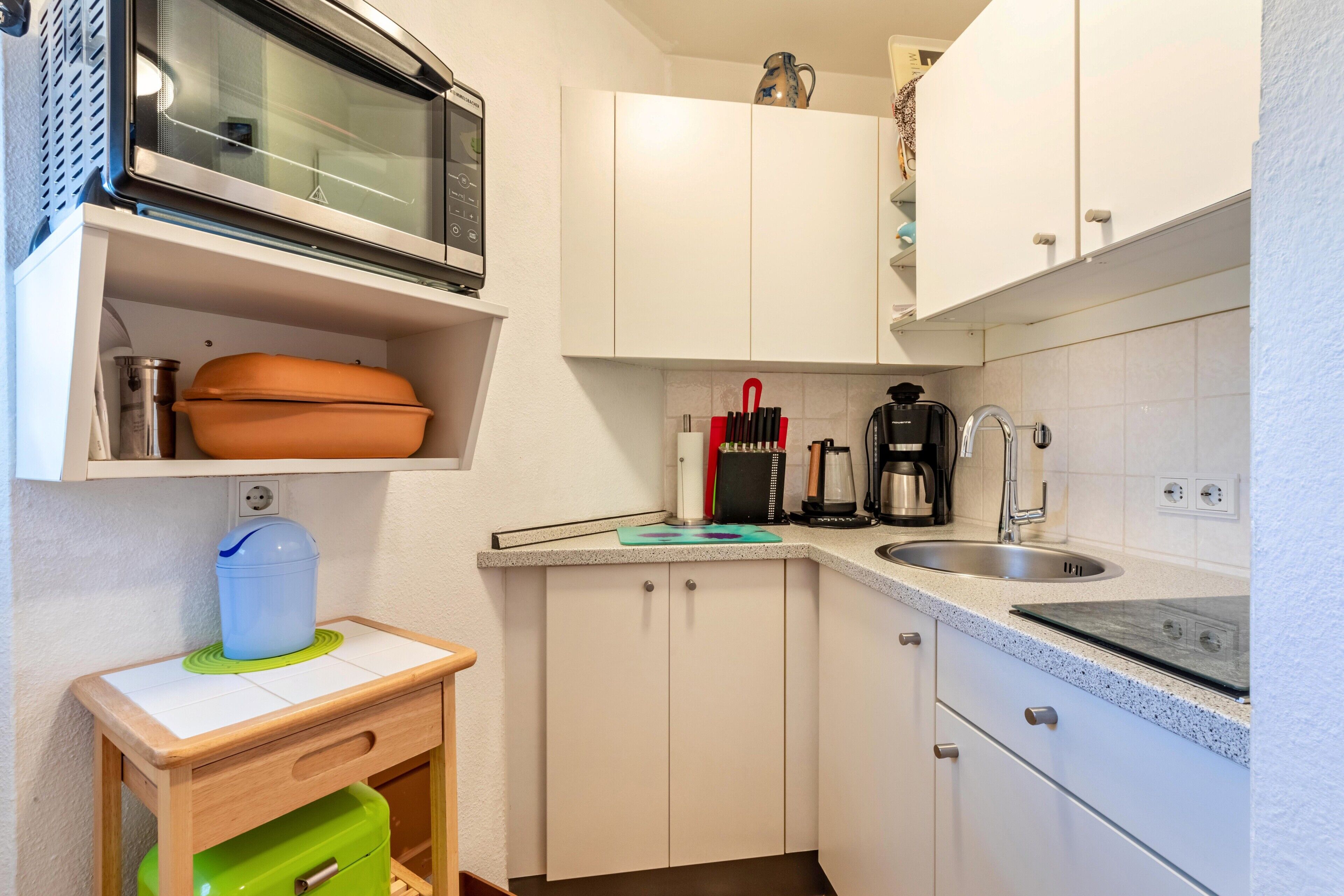 A compact kitchen with a microwave, coffee maker, sink, and a small wooden table with a water container.