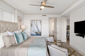 A bedroom with a large bed, a ceiling fan, a painting, and a TV on the wall.