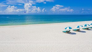 Een strand met wit zand en helderblauw water. Er staan verschillende blauw-witte strandparasols en ligstoelen.