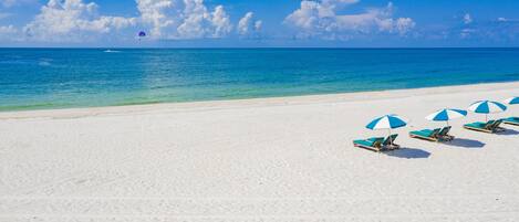 Una playa con arena blanca y agua azul cristalina. Varias sombrillas y sillas de playa en azul y blanco.