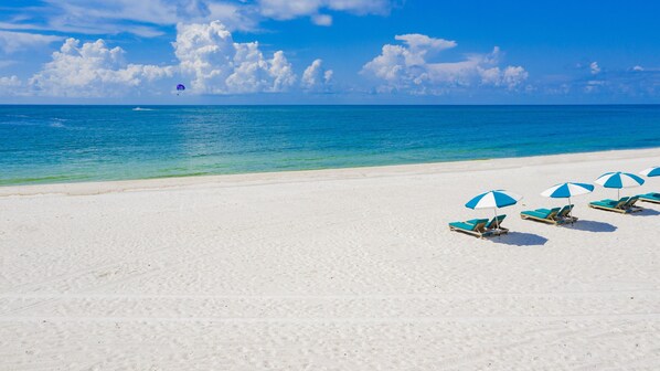 A beach with white sand and clear blue water. There are several blue and white beach umbrellas and lounge chairs.