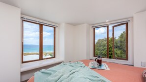 A bedroom with two large windows offering ocean and forest views, a bed with a teal blanket, and a bedside table with a cup and saucer. - Santorini style, luxury apartment, stunning views! (Bowentown)