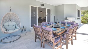 Een buitenontbijtplaats met een ronde tafel voor zes personen, omringd door houten stoelen met gestreepte kussens. Een hangstoel van riet is aan de linkerkant geplaatst, en een glazen schuindeur leidt naar een aangrenzende kamer.