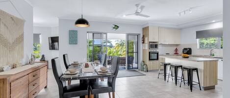 A modern kitchen with a dining area, featuring a large island, bar stools, and a view through the glass doors.
