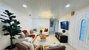 Ett rymligt vardagsrum med ett vitt tak, en stor växt, en soffgrupp, ett soffbord och en TV som är monterad på väggen.