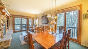 A spacious dining area with a large wooden table and chairs, a stone fireplace, and large windows offering a view of the outdoors. - Walk to Ski, Large 2 Bed 2 Bath Near Lifts 459 (Mammoth Lakes)