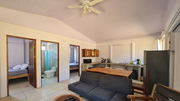 A compact living space with a sofa, a dining table, a wooden coffee table, a ceiling fan, a kitchen area with cabinets and appliances, and a bathroom with a shower curtain. - Una escapada de vacaciones espaciosa y privada en Marbella. (¡Admite mascotas!)  (Marbella)