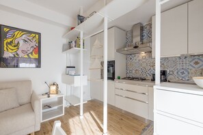 A modern kitchen with white cabinets, a gas stove, and a patterned backsplash. There is a white sofa and a small table with a lamp in the living area.