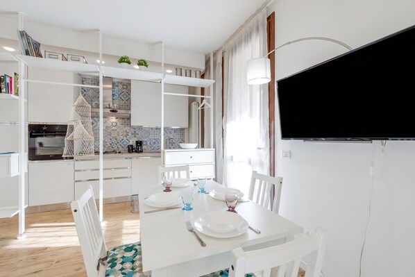 A modern kitchen with white cabinetry, a dining table set for two, and a flat-screen TV mounted on the wall.