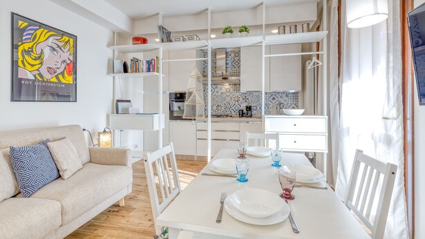 Un salon moderne et bien éclairé, avec un coin repas, des meubles blancs et une cuisine en arrière-plan.