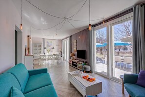 Un soggiorno moderno con divano, tavolino e televisione. C'è un'area pranzo con sedie e tavolo. La stanza ha ampie finestre e vista sull'esterno.