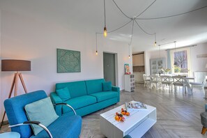 A modern living room with a teal sofa, a white coffee table, and a dining area in the background.