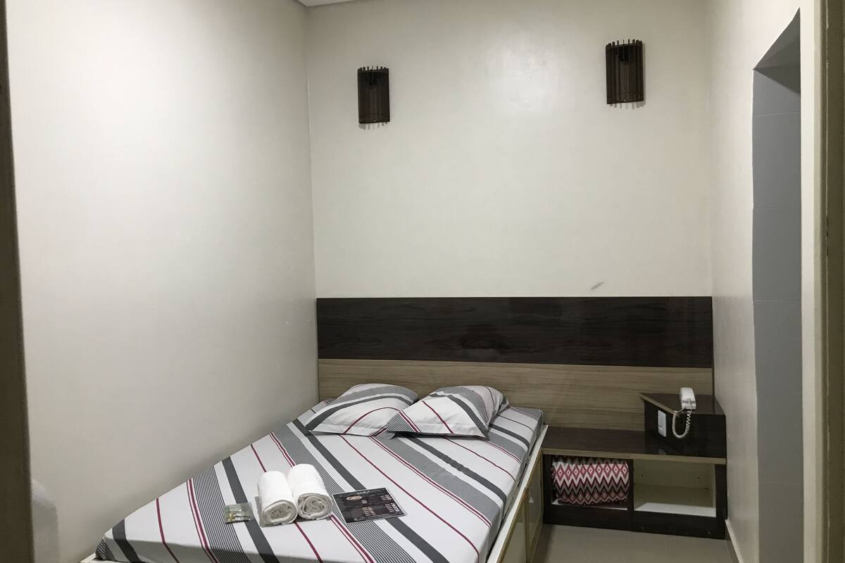 Ein kleines Zimmer mit einem Einzelbett, einer gestreiften Bettdecke, zwei Kissen, einem Nachttisch mit Telefon und einem kleinen Regal.