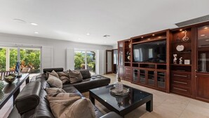 A modern living room with a leather sofa, a coffee table, and a TV. There is a wooden cabinet with glass doors and a large window providing a view of greenery outside. - Jupiter waterfront - dock and new  jacuzzi spa , 3 b 2 b fits 30 “ boat   (Jupiter)