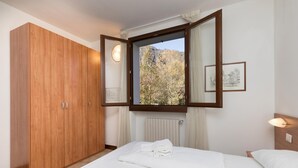 A bedroom with a wooden wardrobe, a bed with white linens, a window with a view of trees and mountains, and a wall-mounted light fixture. - Porto Letizia Rossini 7, Porlezza, Italy (Porlezza)