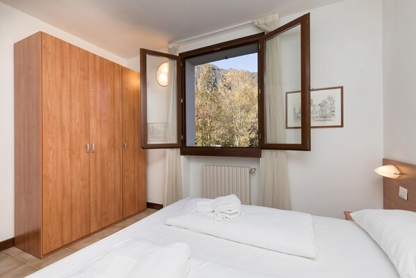 Een slaapkamer met een houten garderobe, een bed met witte lakens, een raam met uitzicht op bomen en bergen, en een wandlamp.