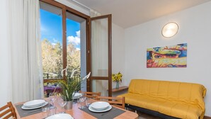 A dining area with a table set for four, a yellow sofa, a painting on the wall, and a window with a view of trees. - Porto Letizia Rossini 7, Porlezza, Italy (Porlezza)
