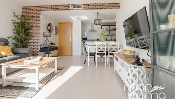 Una sala moderna con una pared de ladrillo decorativa, una zona de comedor con mobiliario blanco y una cocina al fondo.