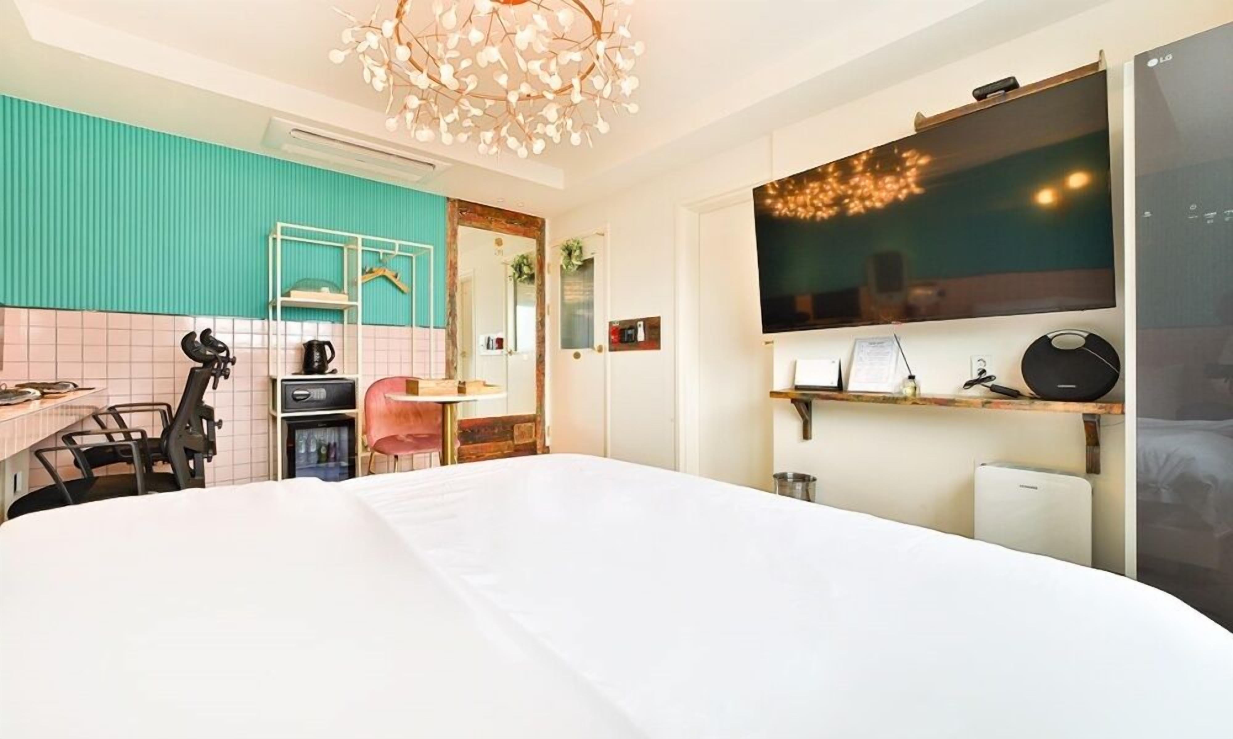 A hotel room with a large bed, a kitchenette with a coffee machine, a desk with a chair, a TV mounted on the wall, and a ceiling light fixture.