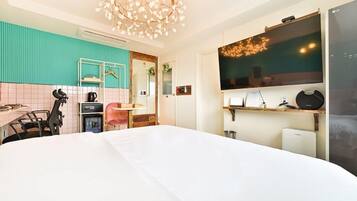 A hotel room with a large bed, a kitchenette with a coffee machine, a desk with a chair, a TV mounted on the wall, and a ceiling light fixture.