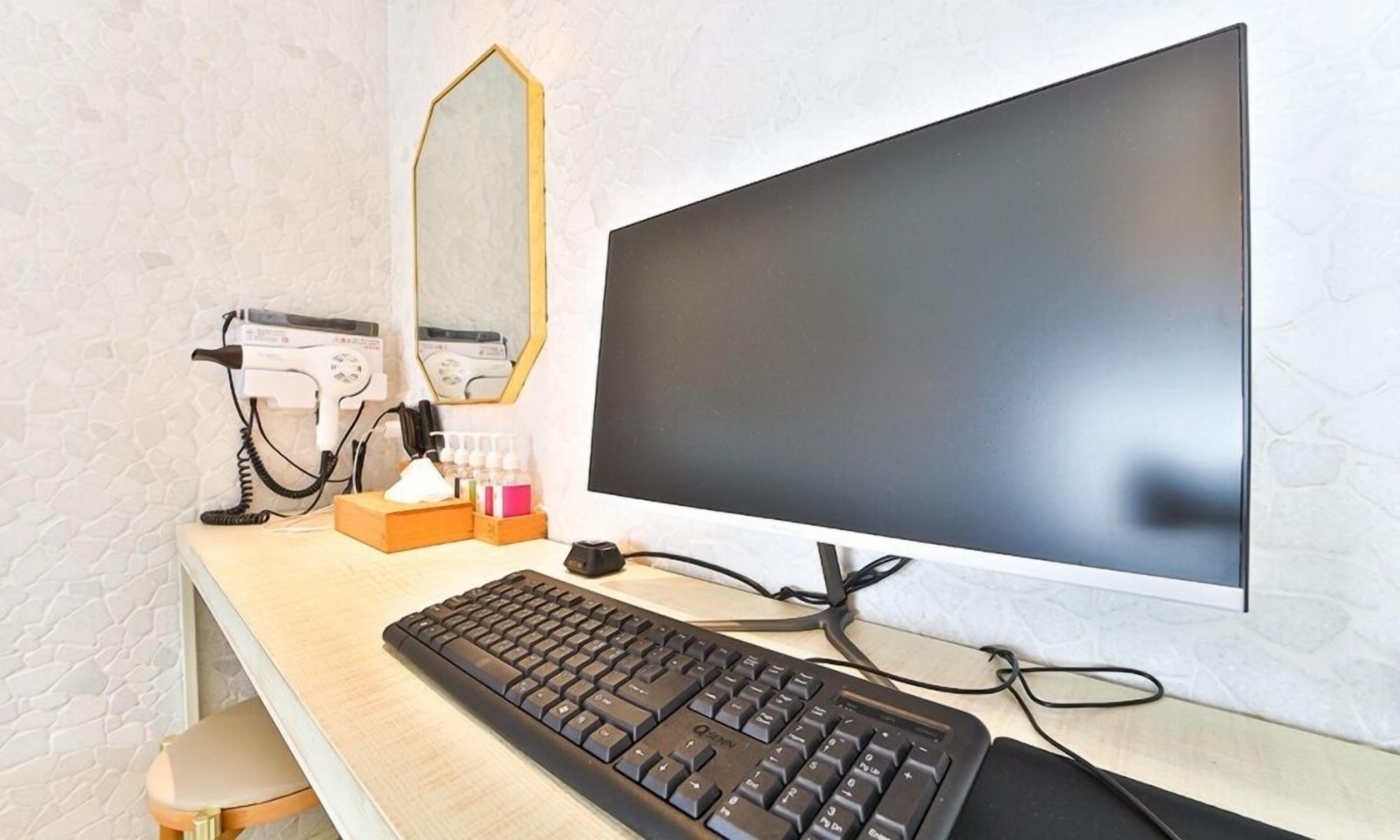 A hotel room with a desk, a desktop computer, a hairdryer, and a mirror.