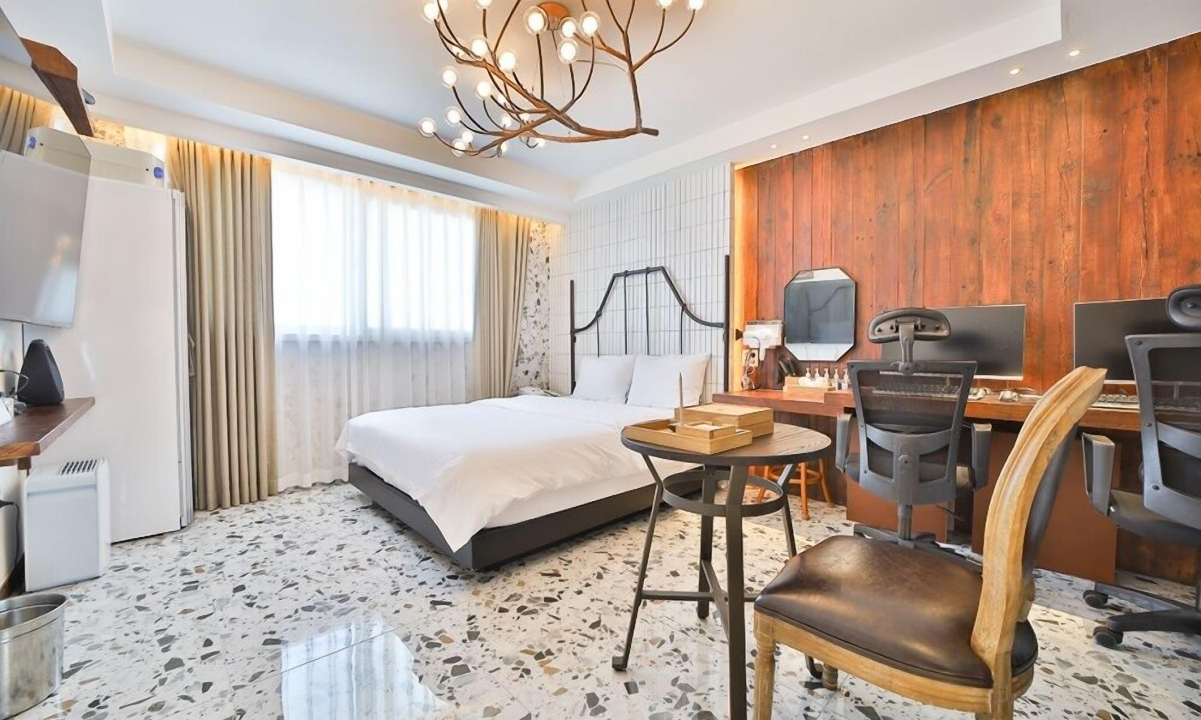 A hotel room with a bed, a desk with a computer, a chair, a mirror, and a unique ceiling light fixture.